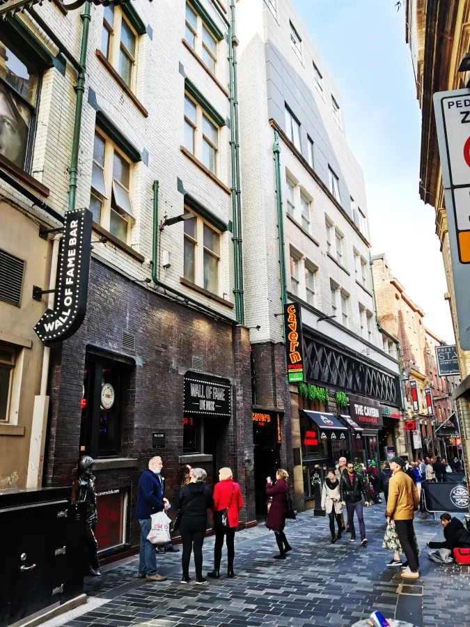 Blick auf eine belebte Straße mit historischen Gebäuden, Cafés und Bars. Menschen stehen vor der 'Wall of Fame' und weiteren Geschäften. Die Architektur ist eine Mischung aus modern und traditionell.