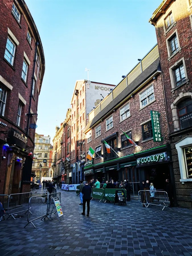Eine belebte Straße mit historischen Gebäuden, auf der McCooley's Bar zu sehen ist. Auf den Gebäuden hängen irische Flaggen und die Straßen sind mit Absperrungen gesäumt.