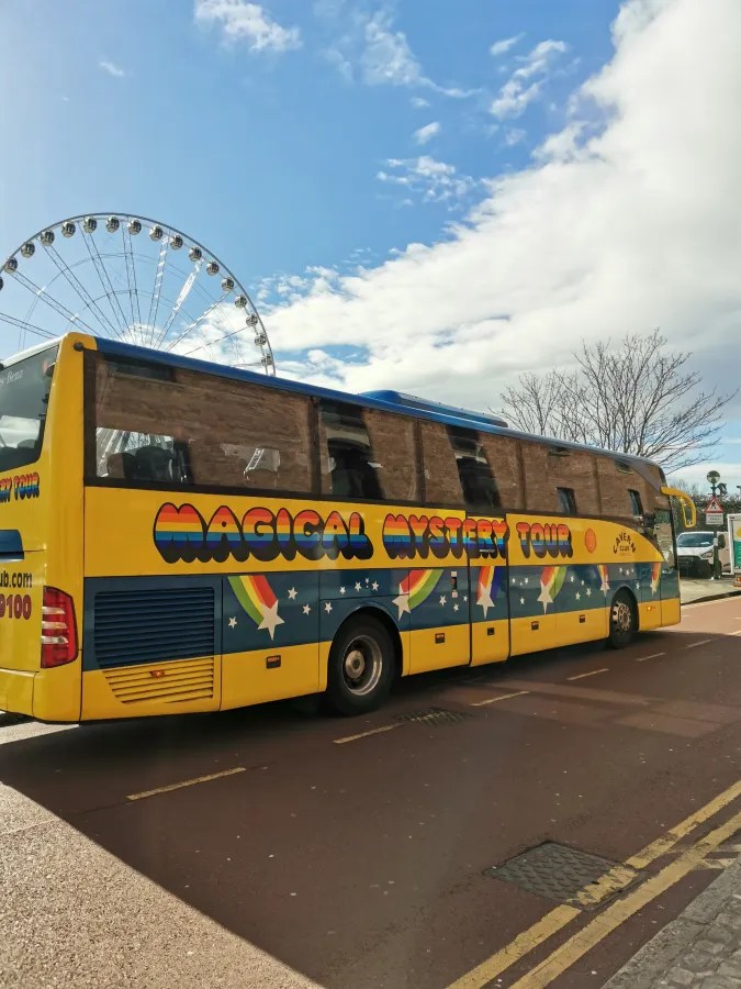 Ein gelber Reisebus mit dem Aufdruck 'Magical Mystery Tour' steht auf einer Straße, im Hintergrund ist ein Riesenrad und ein blauer Himmel mit einigen Wolken zu sehen.
