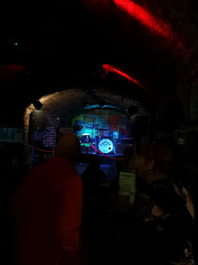 Blick auf die Bühne im Cavern Club, mit Musikern und einem beleuchteten Logo im Hintergrund.