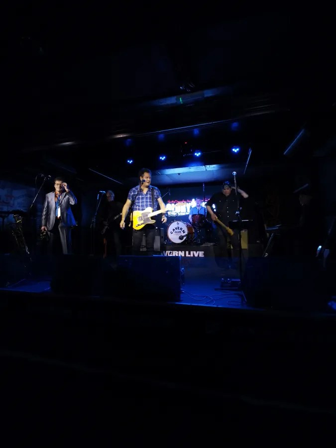 Eine Band spielt auf einer Bühne in einem dunklen Club mit blauen Lichtern, während das Publikum zuschaut.