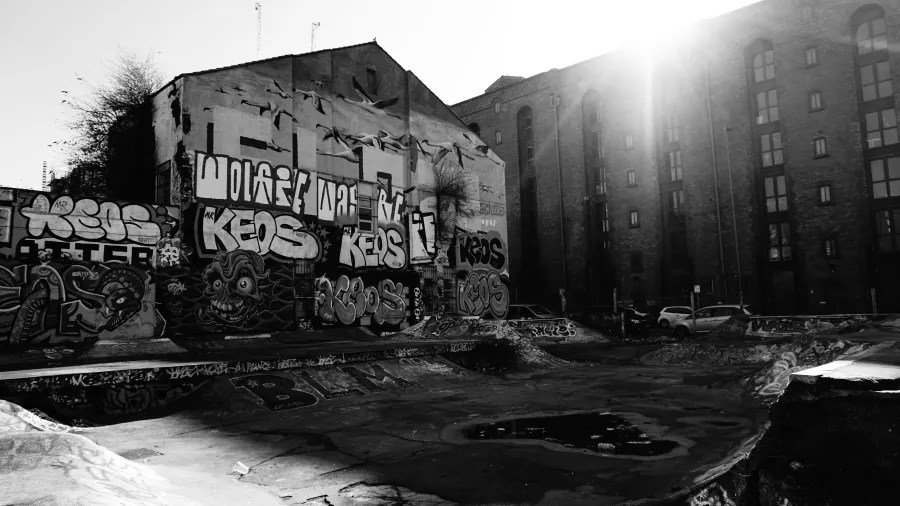 Eine Schwarz-Weiß-Aufnahme eines verlassenen Skateparks mit graffiti-bedeckten Wänden, umgeben von städtischen Gebäuden und Sonnenlicht im Hintergrund.