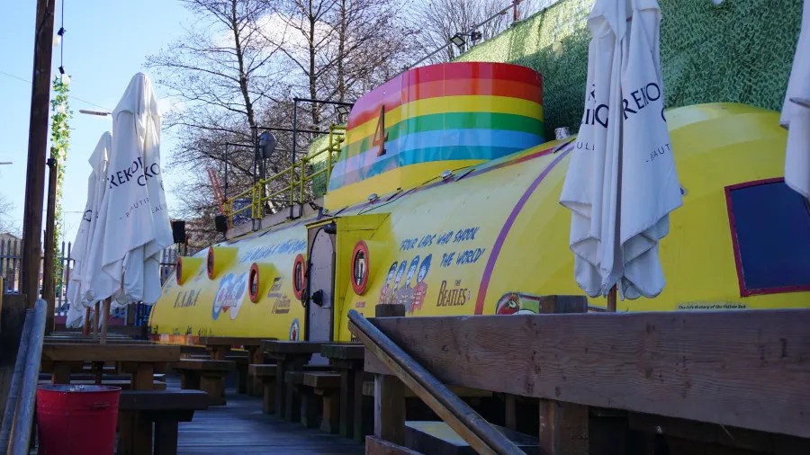 Bunte, gelbe Boot im Stil eines U-Bootes mit Beatles-Motiven, umgeben von Tischen und Sonnenschirmen in einem Außenbereich.