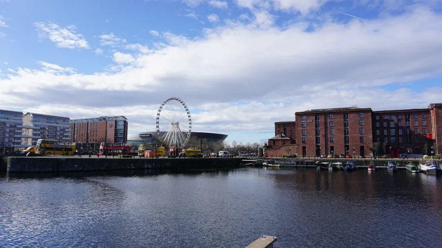 Blick auf einen Hafen mit verschiedenen Gebäuden, Fahrriesenrad und einem Sportstadion im Hintergrund bei klarem Himmel.