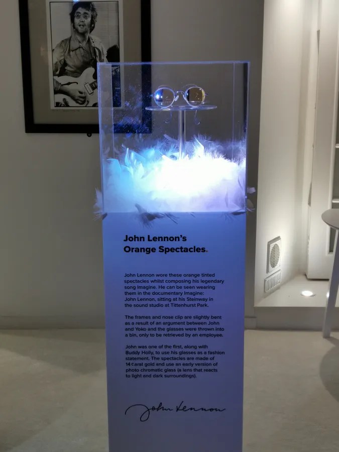 Orangefarbene Brille von John Lennon, ausgestellt in einem Glaskasten auf Federn, mit einer beschreibenden Tafel daneben.