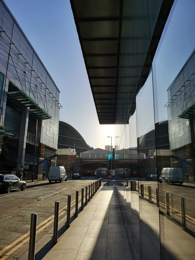 Eine moderne städtische Straße, die von glänzenden Glasfassaden gesäumt ist. Im Hintergrund sind Verkehr und ein Bahnübergang sichtbar, während die Abendsonne lange Schatten wirft.