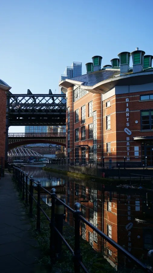 Blick auf einen Kanal mit reflektierenden Ziegelgebäuden und einer Eisenbrücke in einer urbanen Umgebung.