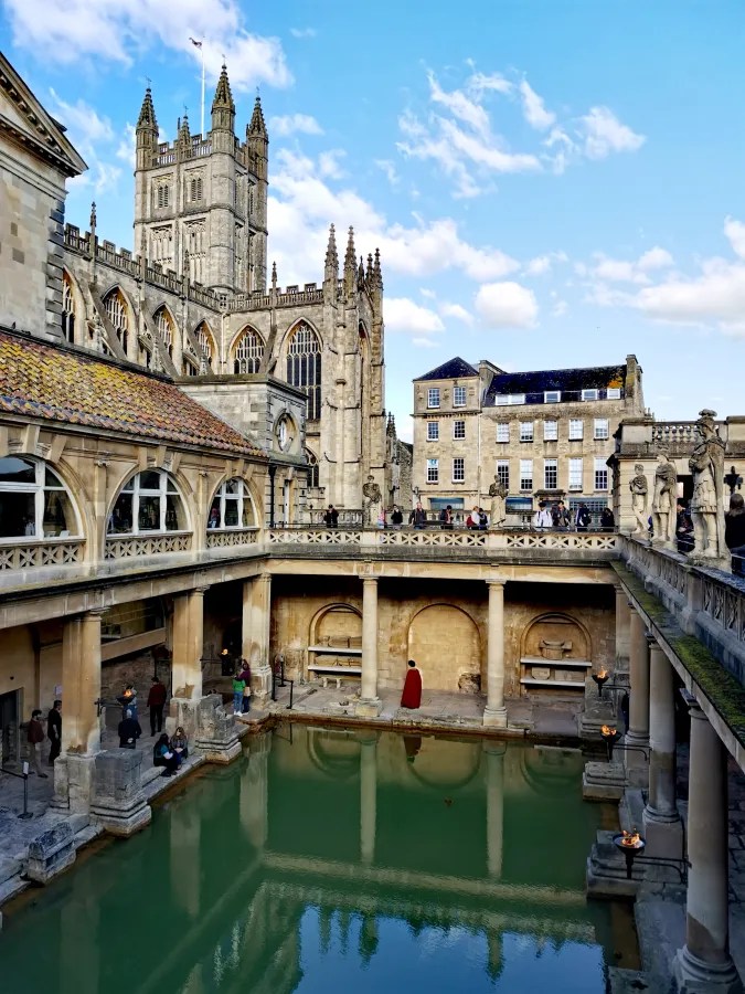 Blick auf die römischen Bäder in Bath, England, mit der beeindruckenden architektonischen Kulisse der Kathedrale im Hintergrund und Besuchern am Wasser.