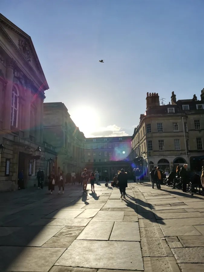Belebter Platz mit Menschen im Sonnenlicht, historische Gebäude und fliegender Vogel im Hintergrund.