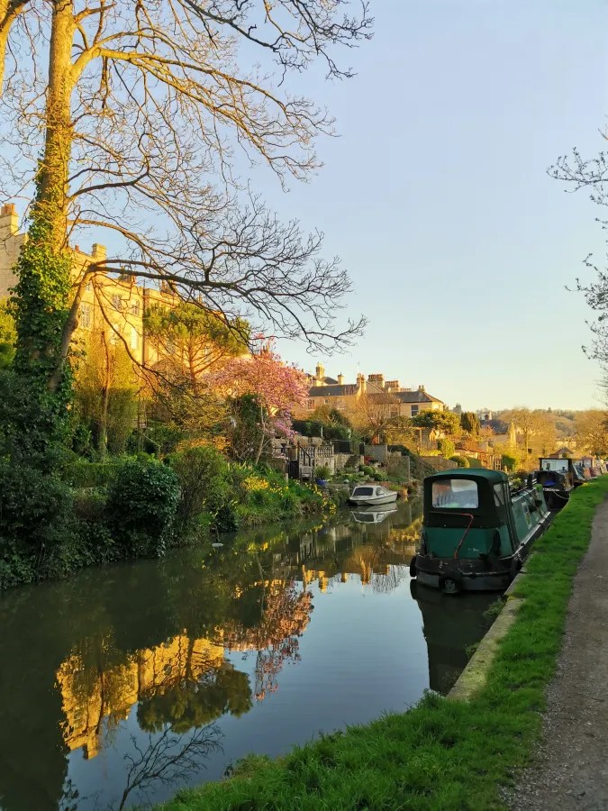 Eine malerische Ansicht eines Kanals mit einem Boot, umgeben von Bäumen und blühenden Pflanzen. Im Hintergrund sind charmante Häuser und ein klarer Himmel zu sehen.