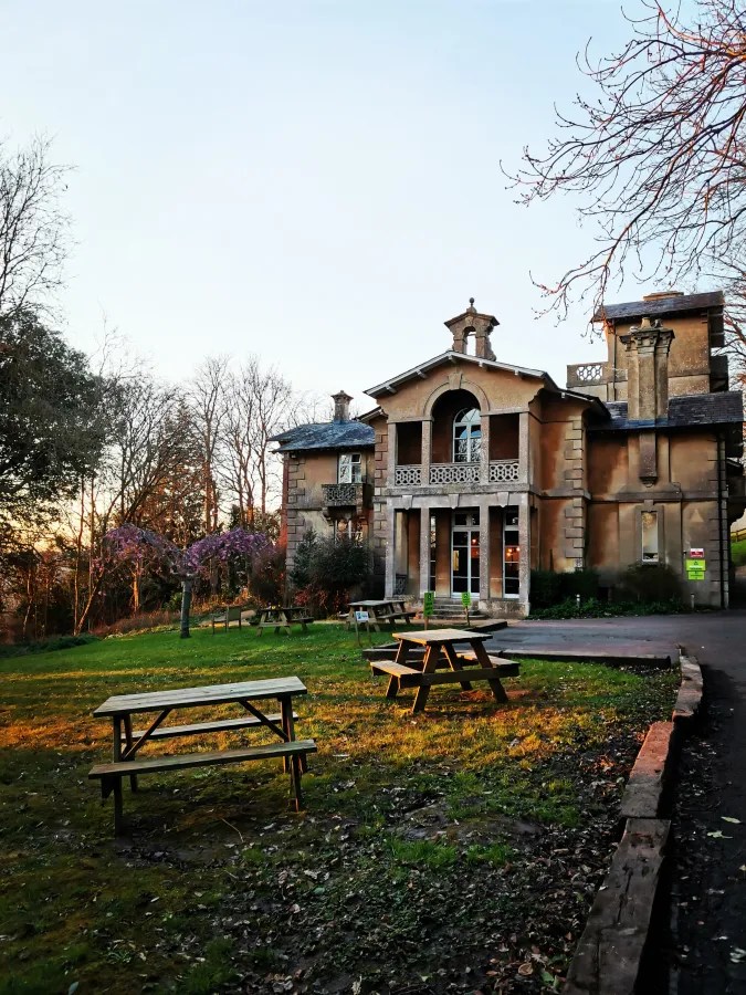 Ein historisches Gebäude mit mehreren Fenstern und einem Balkon, umgeben von einem Garten mit Holzpicknicktischen und Bäumen im Vordergrund.