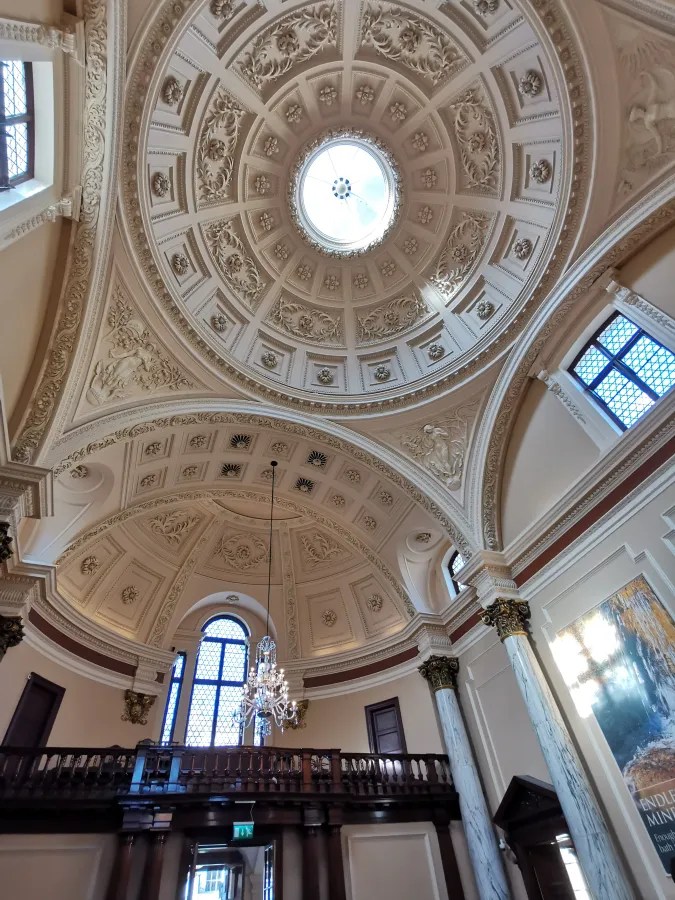 Blick auf eine aufwendig verzierte Decke mit einer zentralen Kuppel und Fenstern, die natürliches Licht hereinlassen.