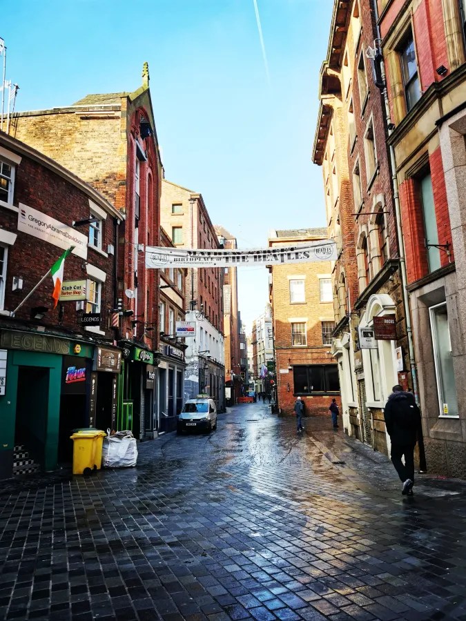 Eine ruhige Straße mit historischen Gebäuden und einem blauen Himmel. Auf der linken Seite sind mehrere Geschäfte, darunter ein Friseursalon. Ein weiteres Geschäft hat eine leuchtende Beschriftung. Ein kleines Auto parkt in der Mitte der Straße, und zwei Personen gehen vorbei. Die Straße hat einen glänzenden Belag, der vom Regen nass aussieht.