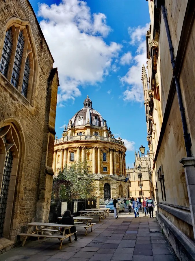 Enge Gasse mit historischen Gebäuden und Blick auf die Kuppel der Bodleian Library in Oxford, unter blauem Himmel mit Wolken.