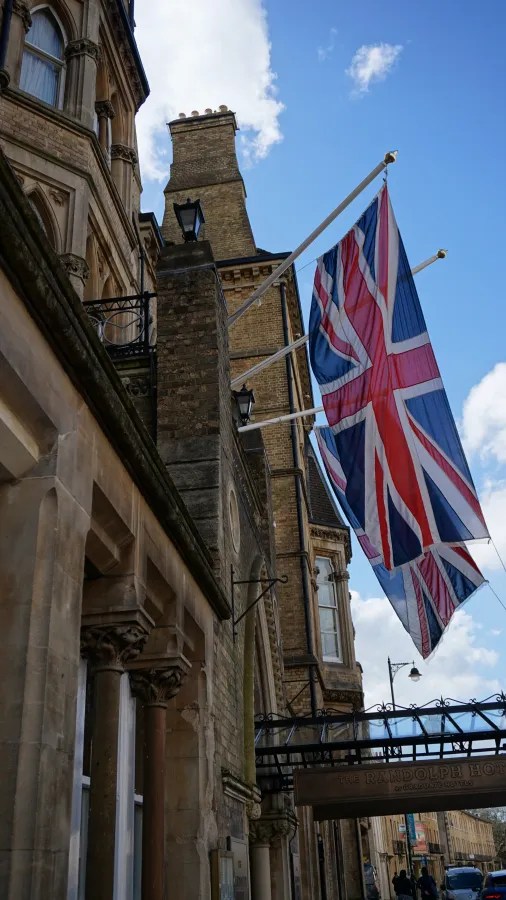 Eine historische Gebäudeansicht mit einer großen britischen Flagge, die über dem Eingang des Randolph Hotels hängt. Der Himmel ist teilweise bewölkt.