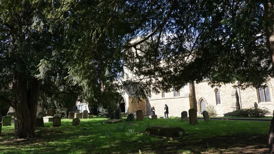 Eine Ansicht eines Friedhofs mit Grabsteinen und Bäumen; im Hintergrund ist eine Kirche zu sehen.