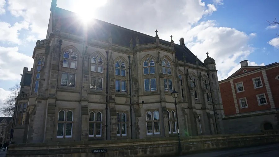 Historisches Gebäude mit gotischen Fenstern, umgeben von niedrigen Bäumen und blauem Himmel, mit Sonnenlicht, das auf die Fassade scheint.