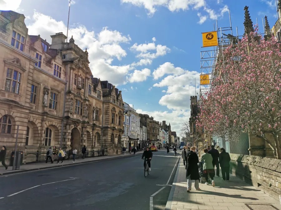 Eine belebte Straße in einer Stadt mit historischen Gebäuden, Radfahrern und Fußgängern. Im Hintergrund sind einige Wolken am Himmel und blühende Bäume zu sehen.
