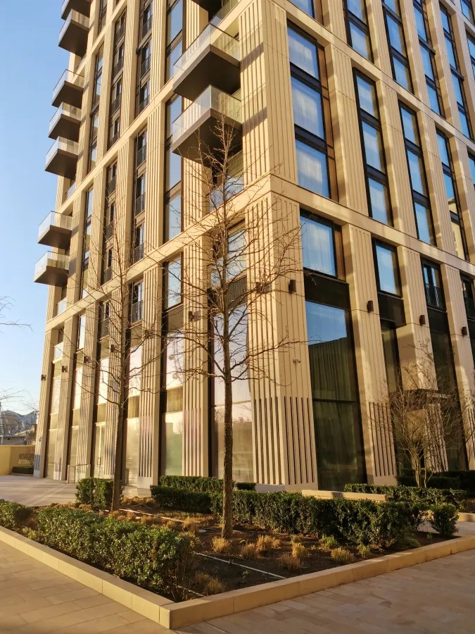 Moderne Architektur eines mehrstöckigen Gebäudes mit großen Fenstern und Balkonen, umgeben von Bäumen und Sträuchern im Außenbereich.