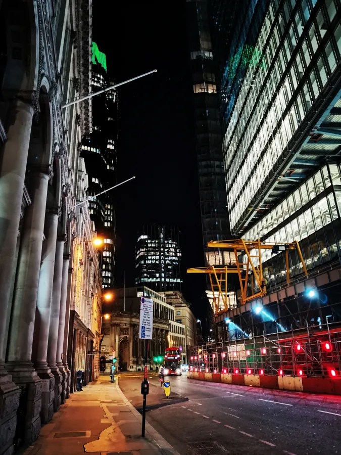 Nachtansicht einer Straße mit modernen Hochhäusern und historischem Gebäude, beleuchtet von Straßenlaternen und Neonlichtern. Ein rotes Bus fährt vorbei, und Baugerüste sind im Bild sichtbar.