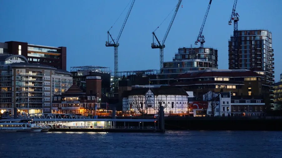 Eine Stadtansicht bei Dämmerung mit modernen Gebäuden und Kränen im Hintergrund, die an einem Fluss liegen.