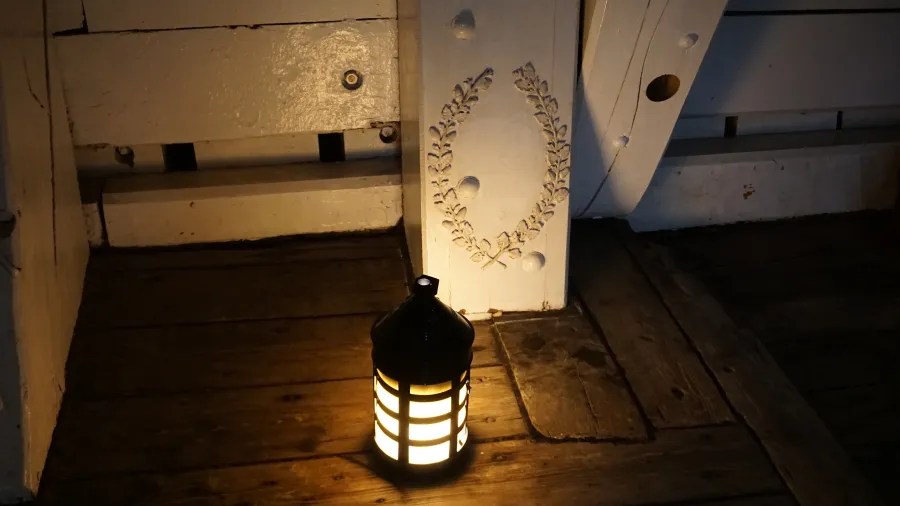 Eine beleuchtete Laterne steht auf einem dunklen Holzboden neben einer weißen Wandverkleidung mit einem dekorativen Muster.