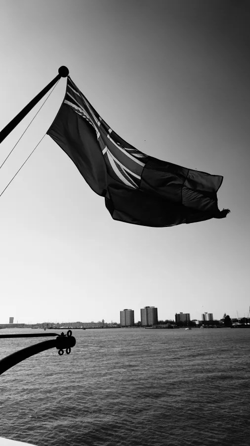 Eine schwarz-weiße Ansicht einer Fahne, die weht, mit einem Stadtbild im Hintergrund und Wasser im Vordergrund.
