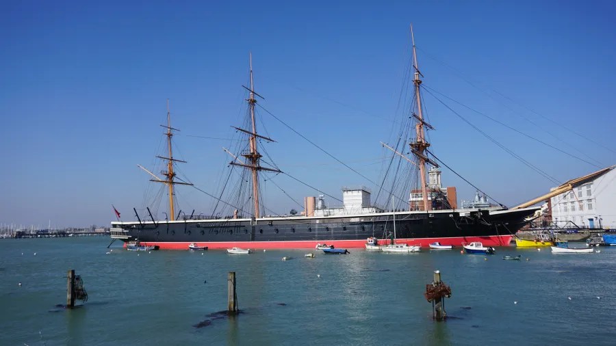 Ein historisches Dampfschiff mit drei Masten im Hafen, umgeben von kleineren Booten und einem klaren blauen Himmel.