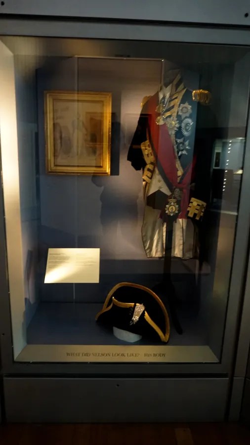 Ein vitrinenpräsentiertes historisches Militäruniformensemble mit Orden, neben einem Bild im Hintergrund und einem Hut im Vordergrund.