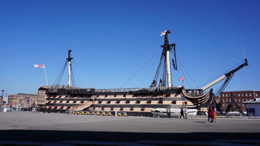 Ein historisches Schiff im Hafen, unter blauem Himmel mit Fahnen. Menschen gehen vorbei.
