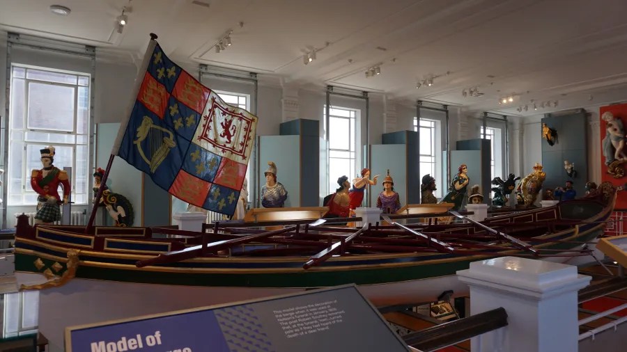 Ein Modell eines historischen Bootes mit einer bunten Flagge und mehreren Figuren in historischen Kostümen im Hintergrund, in einem hell erleuchteten Ausstellungsraum.