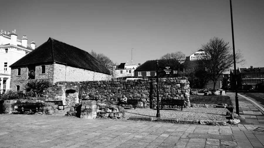 Eine schwarz-weiße Aufnahme eines alten Steinhauses mit einem schrägen Dach, umgeben von einer Steinmauer und Bänken in einer parkähnlichen Umgebung.