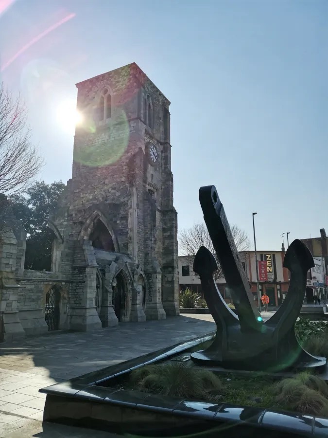 Eine historische Kirche mit einem Glockenturm im Vordergrund, neben einem großen Anker in einem städtischen Platz bei sonnigem Wetter.