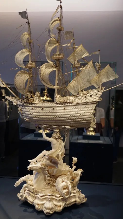 Detailansicht eines kunstvoll gefertigten Schiffsmodells aus Elfenbein, das auf einer dramatischen Basis mit einer Figur sitzt, die eine mythologische Gestalt darstellt. Das Schiff hat mehrere Segel und ist aufwendig verziert.