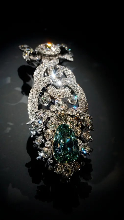 Eine detaillierte Brosche mit glitzernden Diamanten und einem großen grünen Edelstein in Tropfenform, auf einem dunklen Hintergrund.