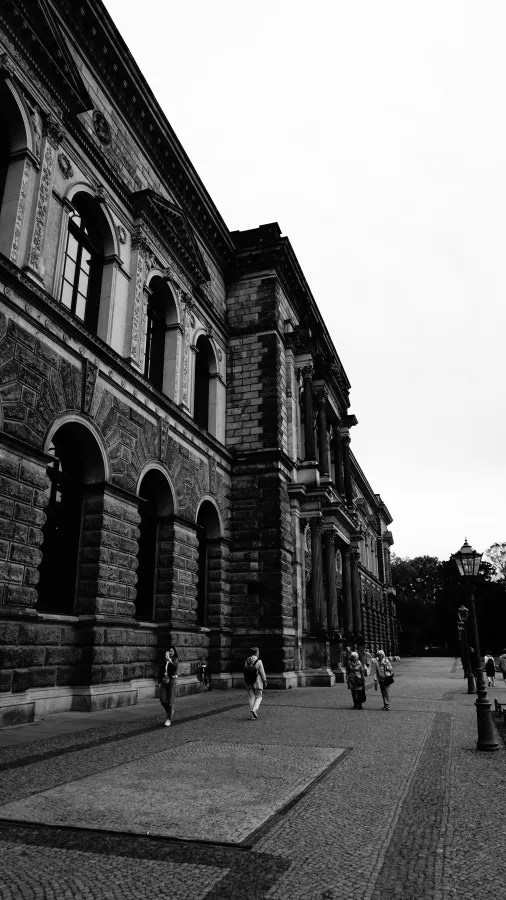 Ein historisches Gebäude mit einer beeindruckenden Fassade, gesehen aus einem niedrigen Winkel. Menschen gehen auf einem gepflasterten Platz vorbei, während der Himmel übercast ist.