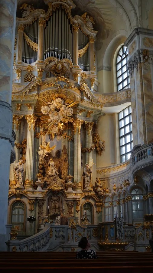 Innenansicht einer Kirche mit goldenen Verzierungen, einer Orgel und einem kunstvoll gestalteten Altar. Eine Person sitzt auf einer der Bänke.