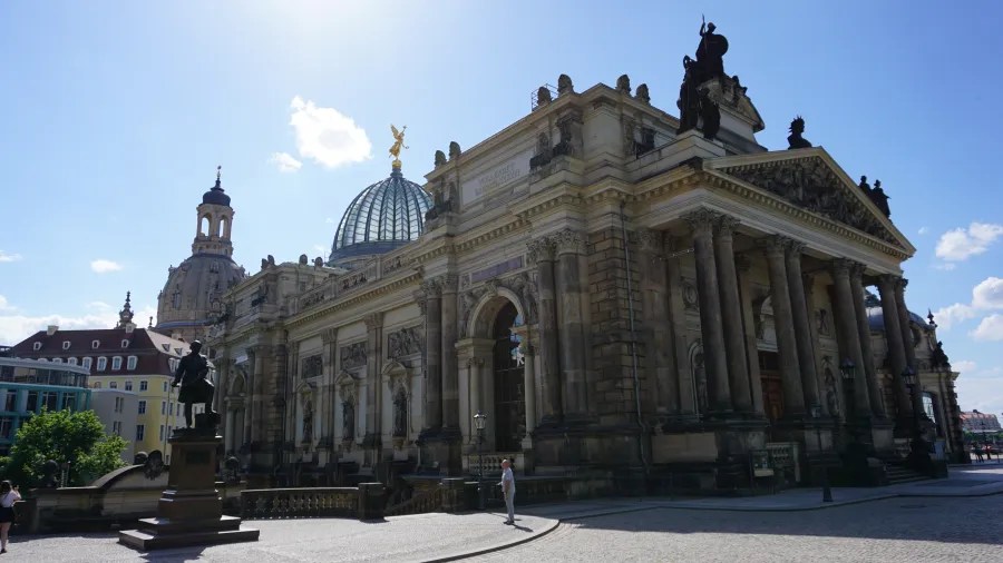 Eine Ansicht des beeindruckenden Zwinger-Palastes in Dresden mit einer Statue im Vordergrund und einer Kuppel im Hintergrund, bei klarem Himmel.