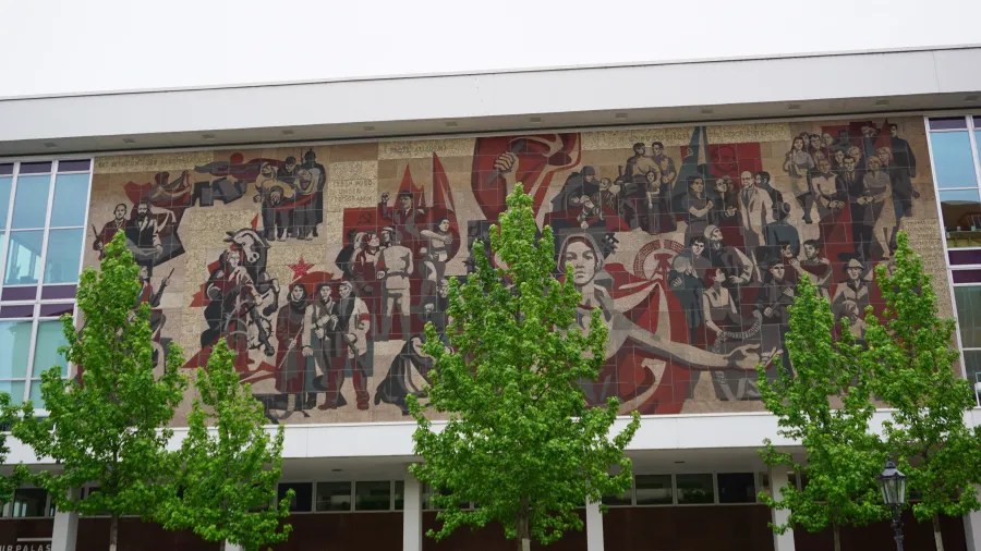 Ein Wandmosaik, das verschiedene Figuren und Szenen darstellt, darunter historische und kulturelle Elemente, umgeben von grünen Bäumen.