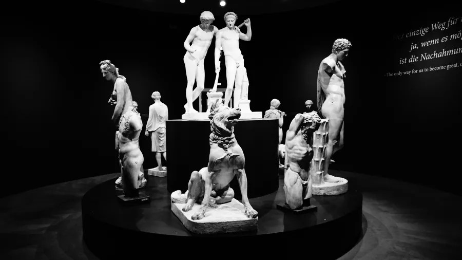 Eine Vielzahl von skulpturalen Statuen in einem Ausstellungsraum, darunter menschliche Figuren und ein Hund, beleuchtet von einer dunklen Umgebung.