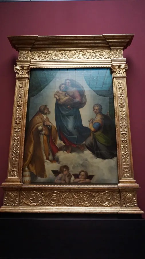 Ein Gemälde von Engeln, die auf einer Wolke liegen, während ein Mann und eine Frau, mit einer weiteren Figur in der Mitte, unter einem Himmel mit blauen Tönen dargestellt sind. Der Holzrahmen ist reich verziert.