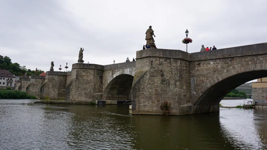 Eine Steinbrücke mit Statuen auf den Pfeilern, die über einen Fluss führt, umgeben von einer malerischen Landschaft und einem bewölkten Himmel.