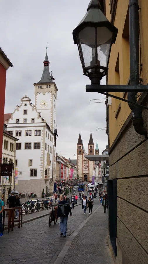 Blick auf eine belebte Straße mit historischen Gebäuden, einer Kirche mit zwei Türmen und einem Lampe an der Wand.