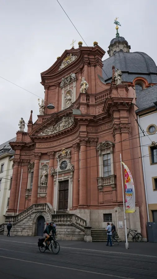 Fassade einer barocken Kirche mit detailreicher Architektur und Statuen, umgeben von Stadtleben.