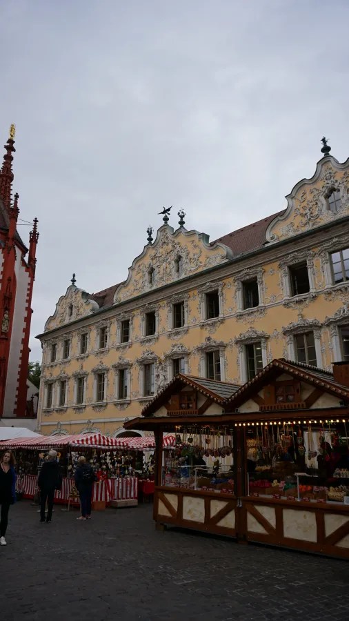 Historisches Gebäude mit barocker Fassade in einer Stadt, umgeben von einem Markt mit bunten Ständen.