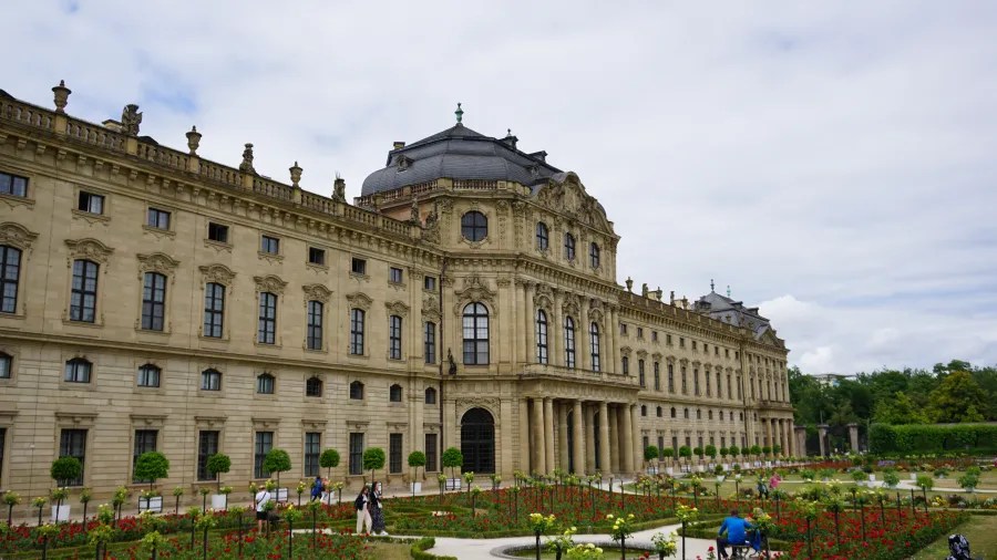 Ein historisches Gebäude mit einer beeindruckenden Fassade und einem angelegten Garten, in dem Menschen spazieren gehen.