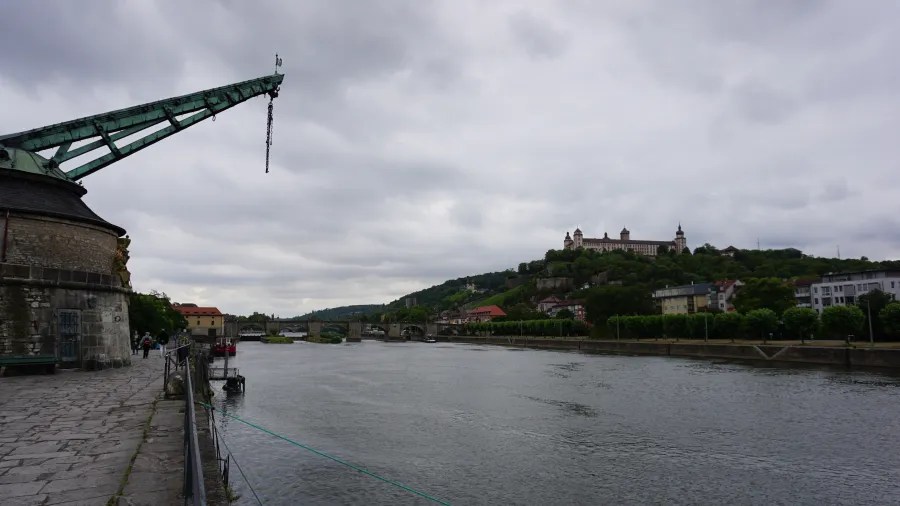Blick auf den Fluss mit einer alten Kränen und einer Burg im Hintergrund, umgeben von grünen Hügeln und bewölkten Himmel.