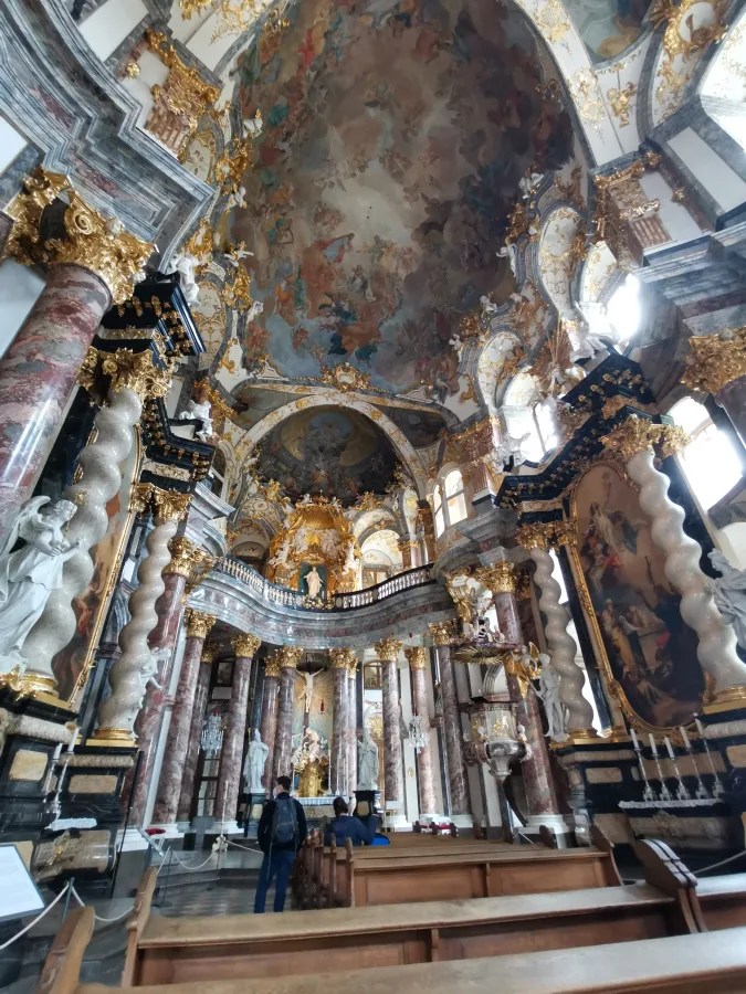 Innenansicht einer prächtig gestalteten Kirche mit goldenen Verzierungen, kunstvoll bemaltem Deckenfresko und ausdrucksvollen Skulpturen, Menschen stehen im Vordergrund.