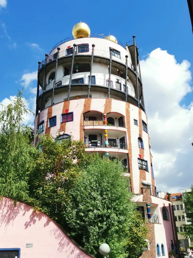 Bunt gestaltetes, rundes Gebäude mit goldenen Kuppeln und grünen Bäumen im Vordergrund unter einem blauen Himmel.
