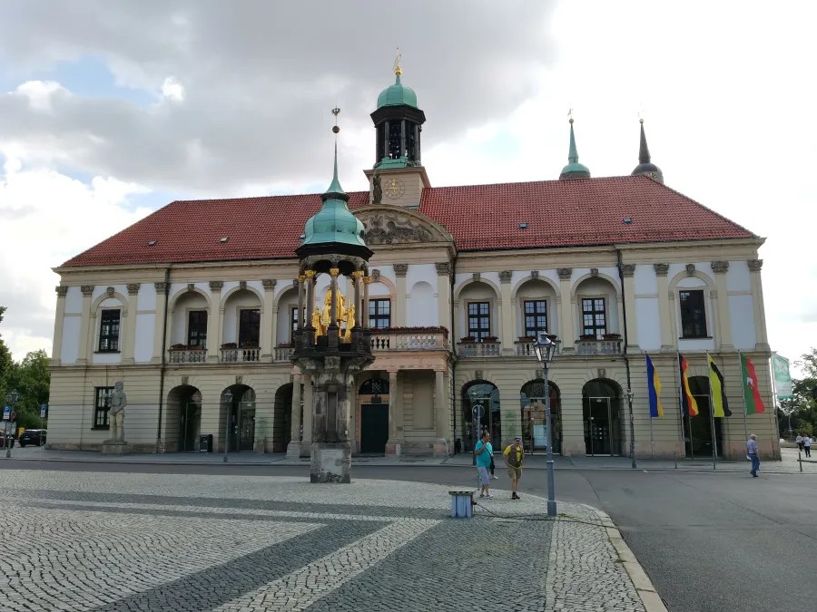 Historisches Gebäude mit rotem Ziegeldach und grünen Kuppeln, umgeben von einem gepflasterten Platz und mehreren Fahnen.